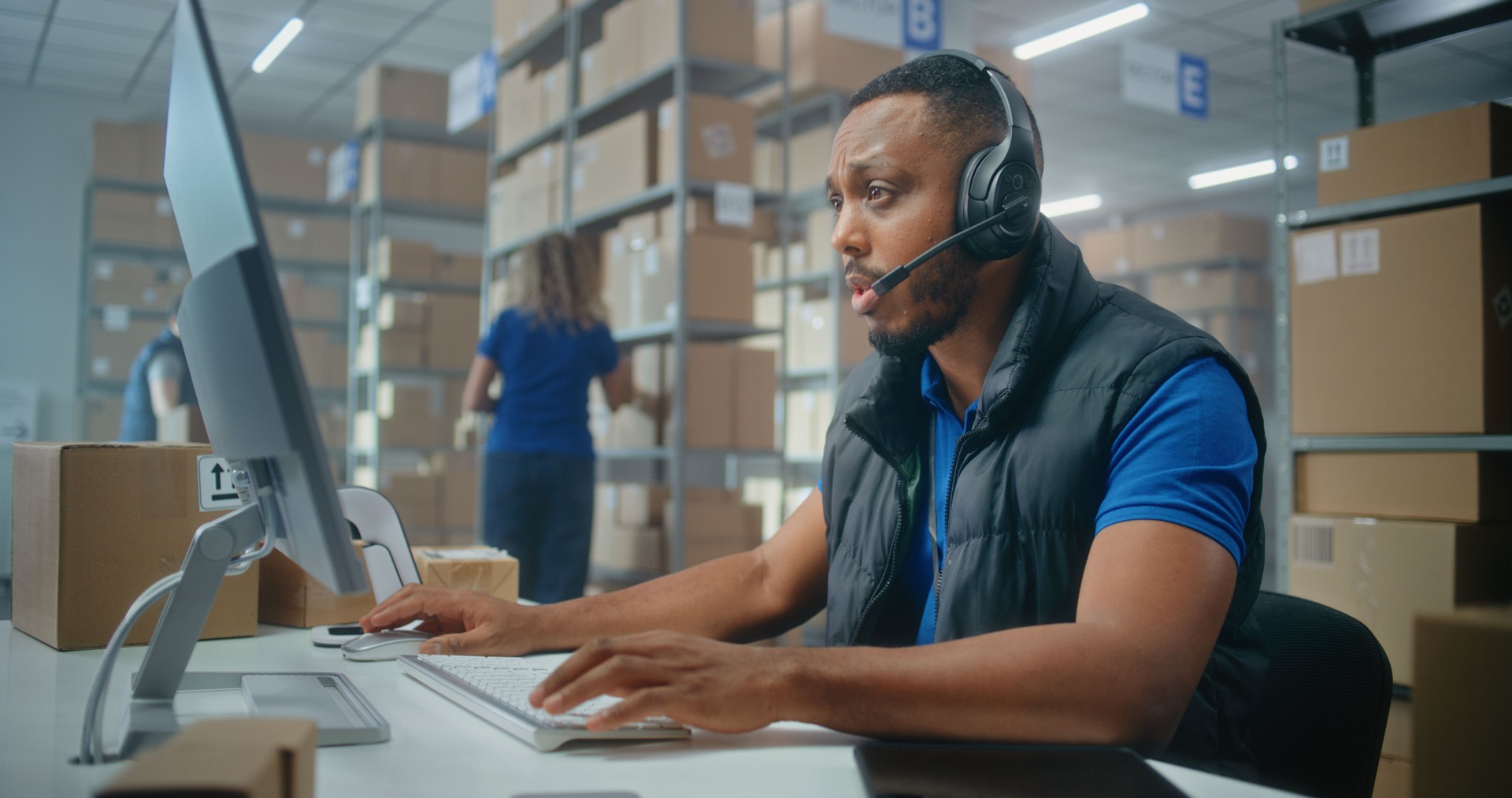African American logistics coordinator wearing headset chats with customer on call, uses computer